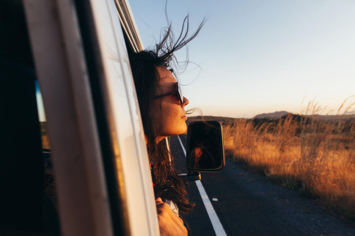 Female with her hair blowing wearing sunglasses hanging out a car window with rural landscape.